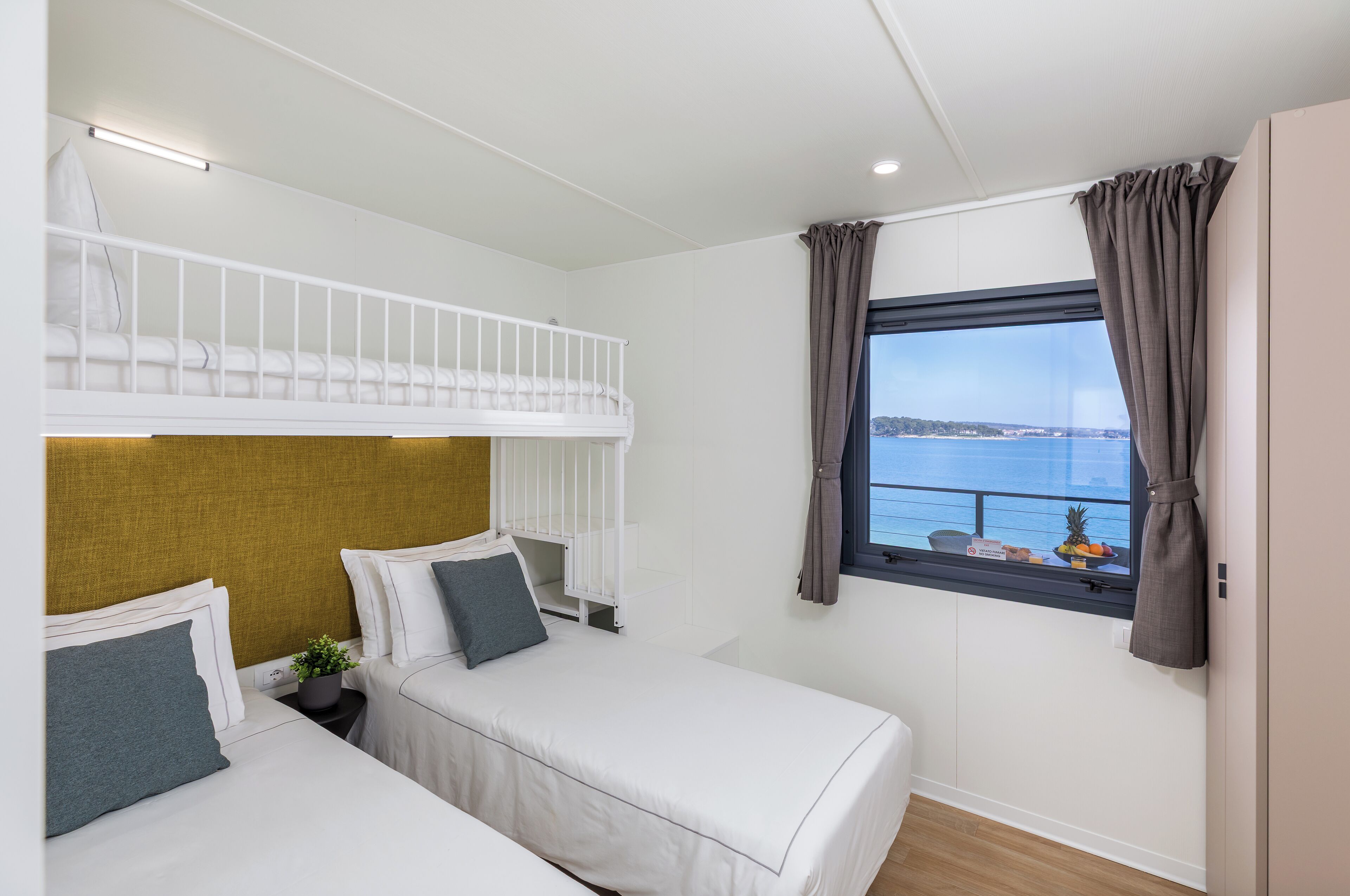 A hotel room with a bunk bed, two single beds, a window with a view of the sea, and a small table with fruit.