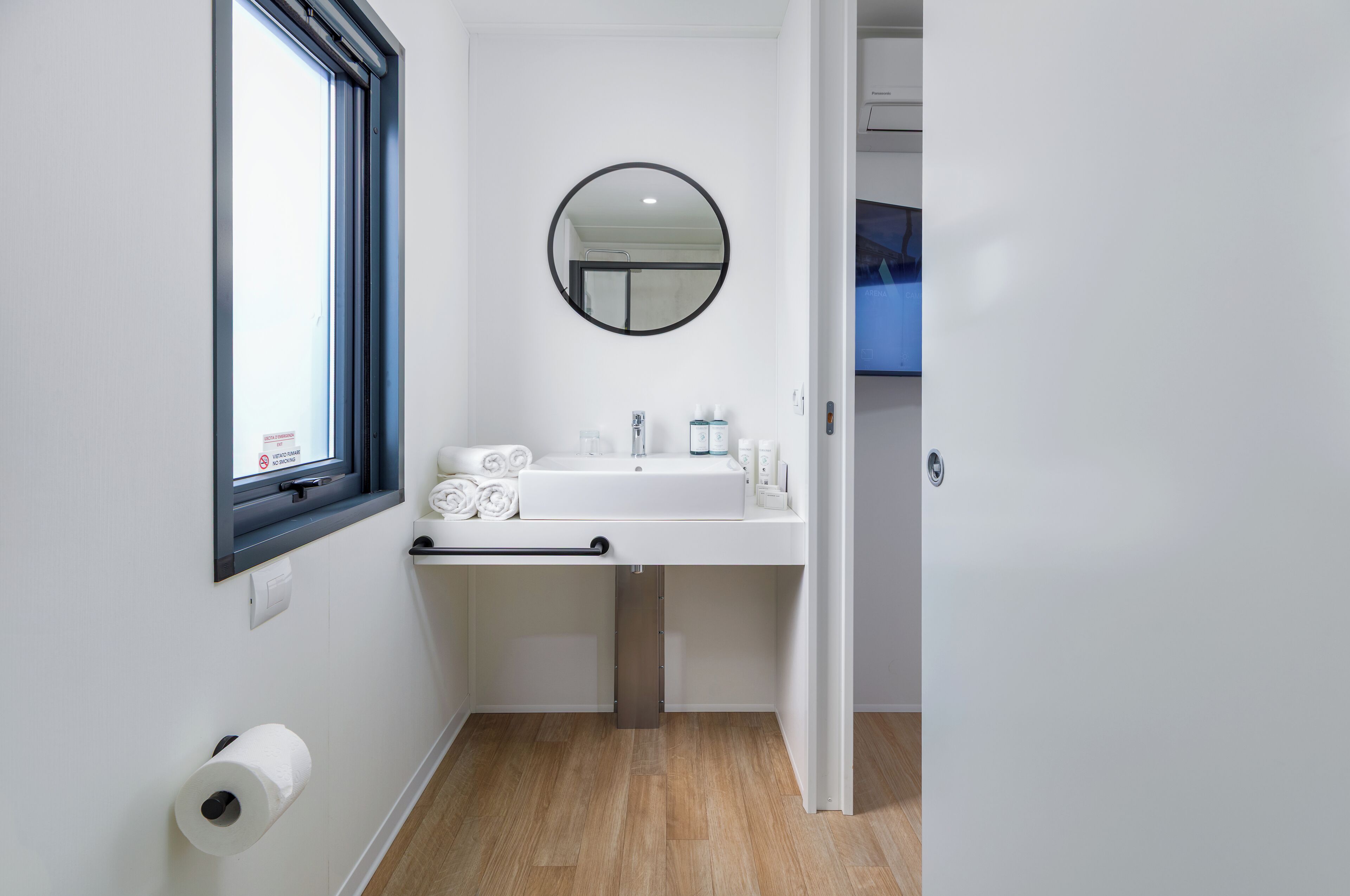Ein Badezimmer mit einem weißen Waschbecken, einem runden Spiegel, einem Toilettenpapierhalter und einem Fenster.