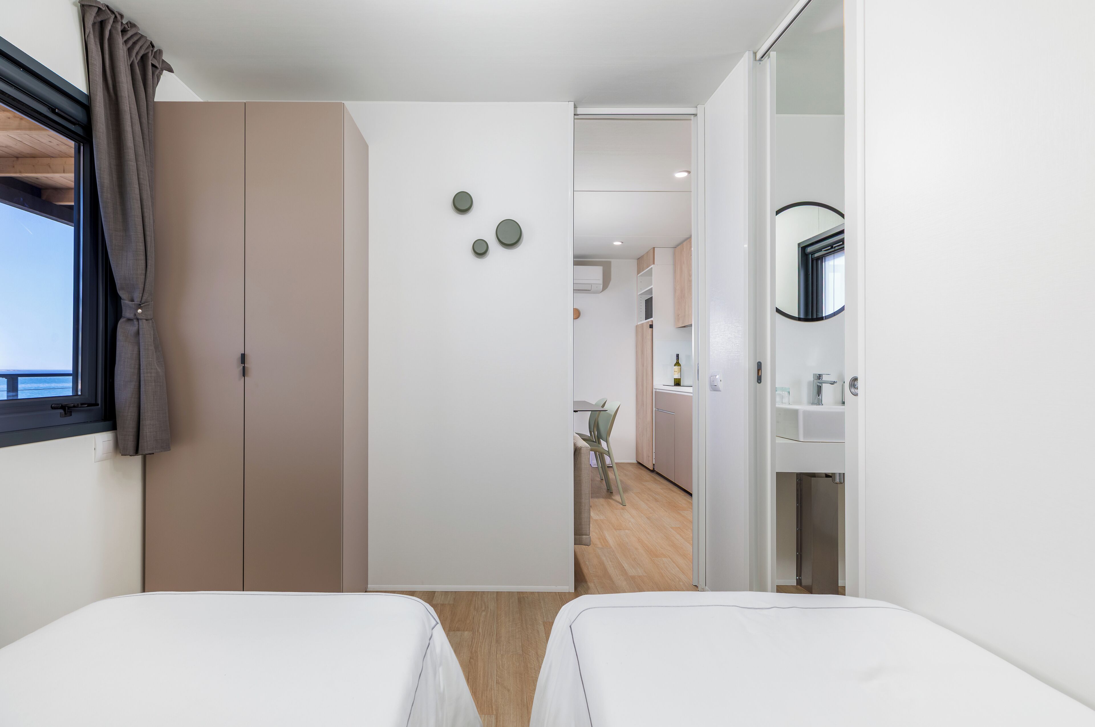 A modern hotel room with two beds, a large wardrobe, and a view of the kitchen area.