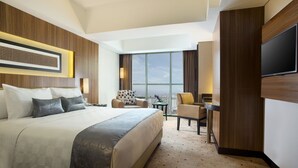 Hotelkamer met een groot bed, twee fauteuils, een kleine tafel, een televisie en een groot raam met uitzicht op de stad.