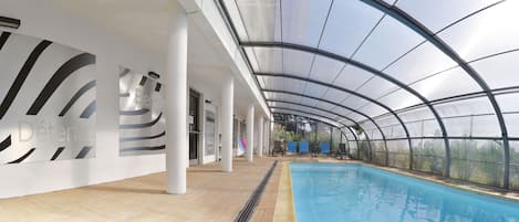 An indoor swimming pool with a glass ceiling, white walls with black wave patterns, and blue chairs along the poolside.