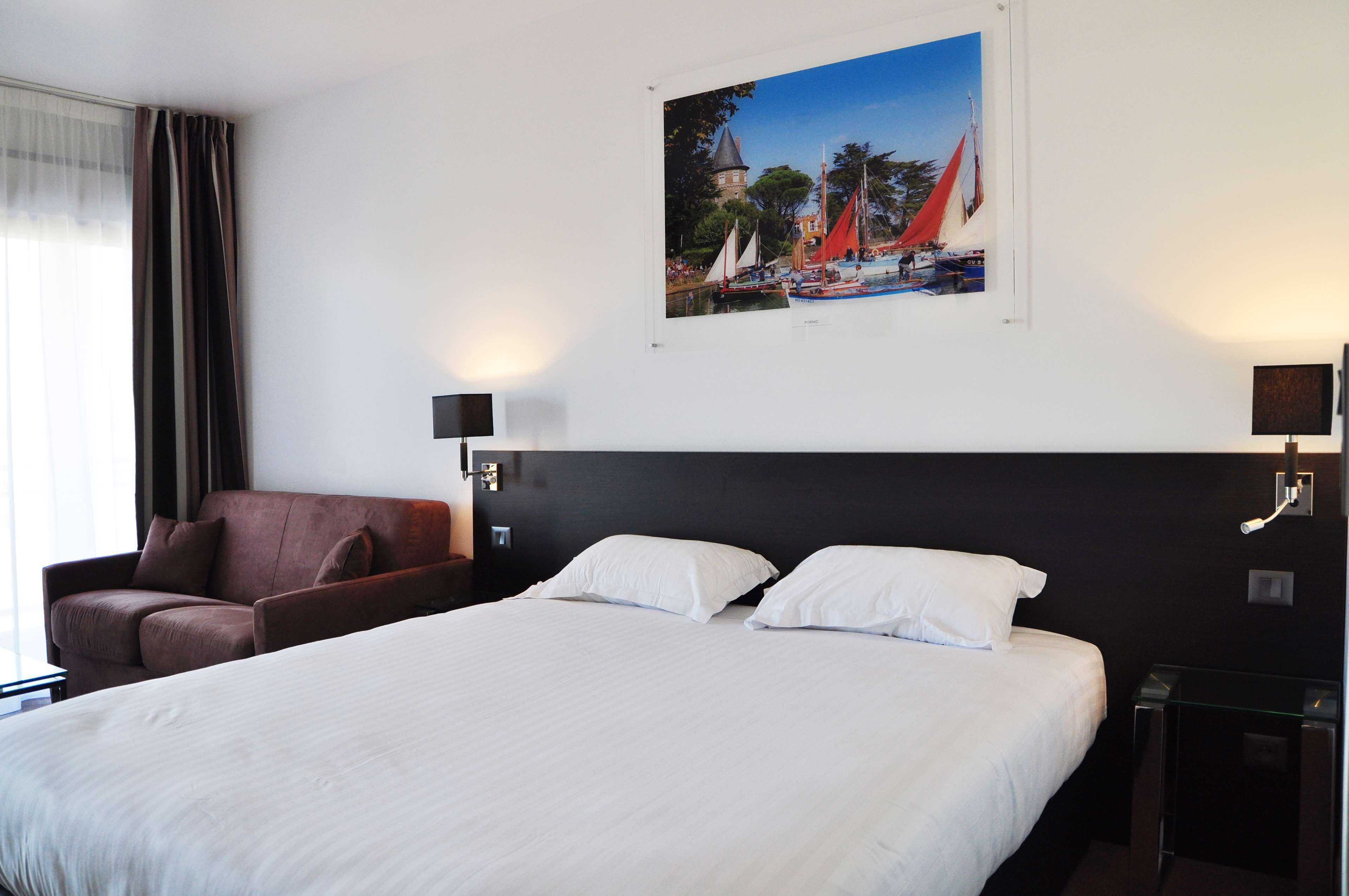 Una camera d'albergo con un letto grande, una poltrona marrone, una TV a parete e un quadro raffigurante una veduta costiera.