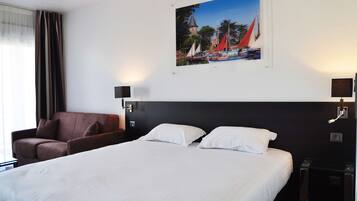 Una camera d'albergo con un letto grande, una poltrona marrone, una TV a parete e un quadro raffigurante una veduta costiera.