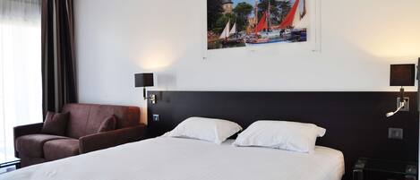 A hotel room with a large bed, a brown armchair, a wall-mounted TV, and a painting of a coastal scene.