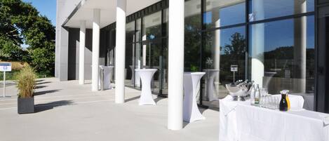A modern building with large windows and a covered outdoor area with white tables and chairs.