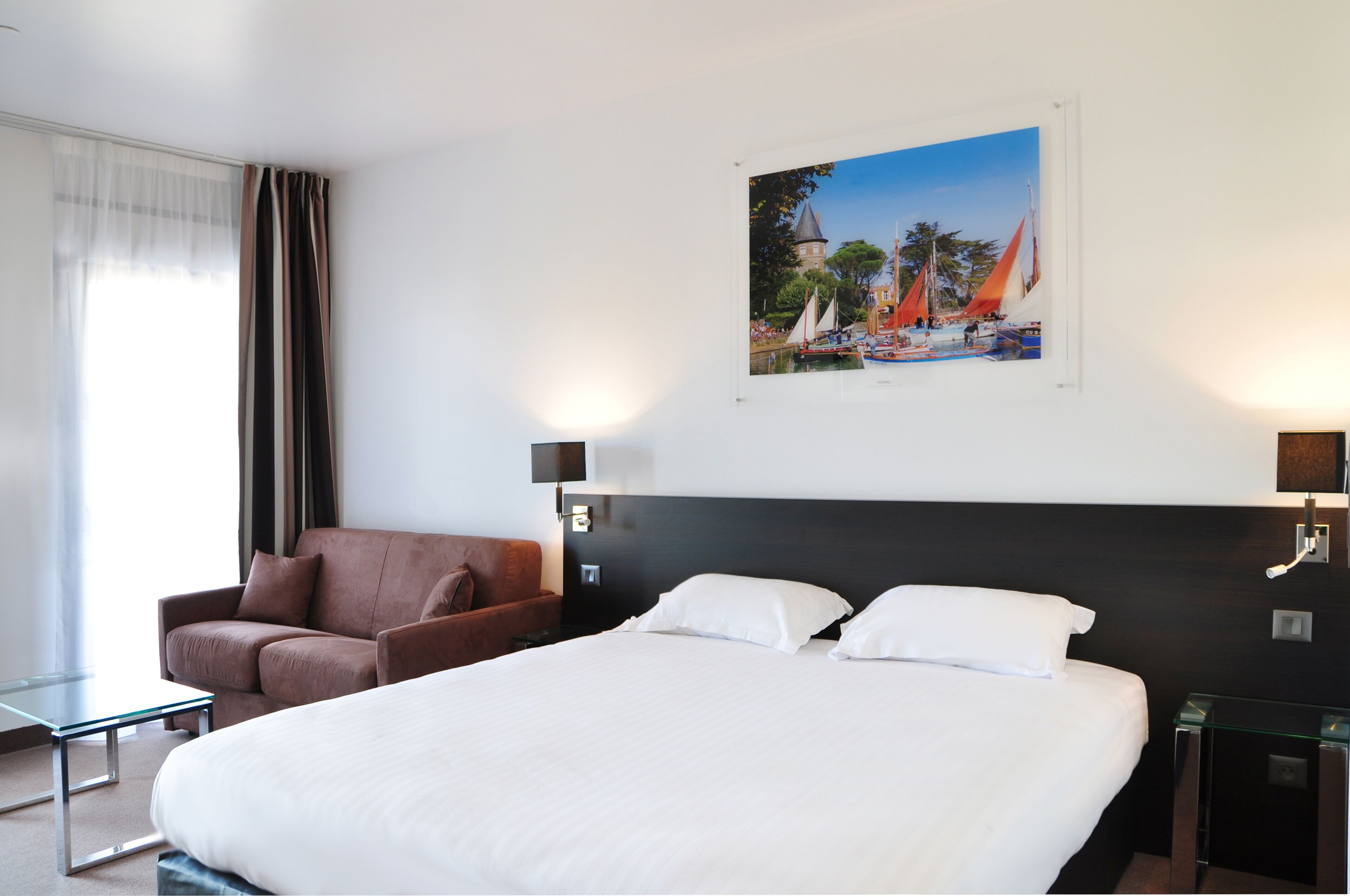 A hotel room with a large bed, a brown armchair, a glass coffee table, and a painting of a coastal scene on the wall.