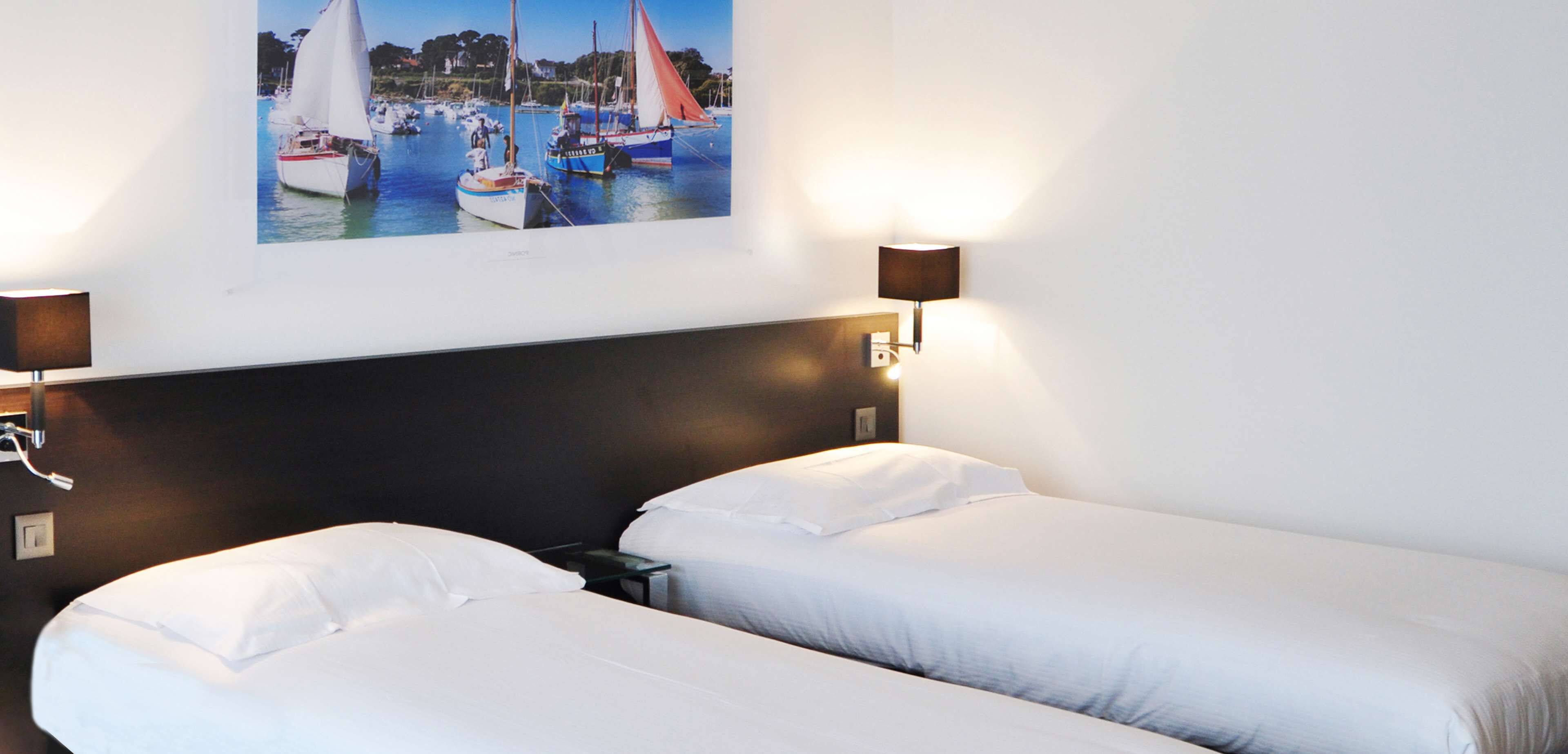 Una habitación de hotel con dos camas individuales, un cabecero oscuro, un cuadro de veleros en la pared y dos lámparas de pared.