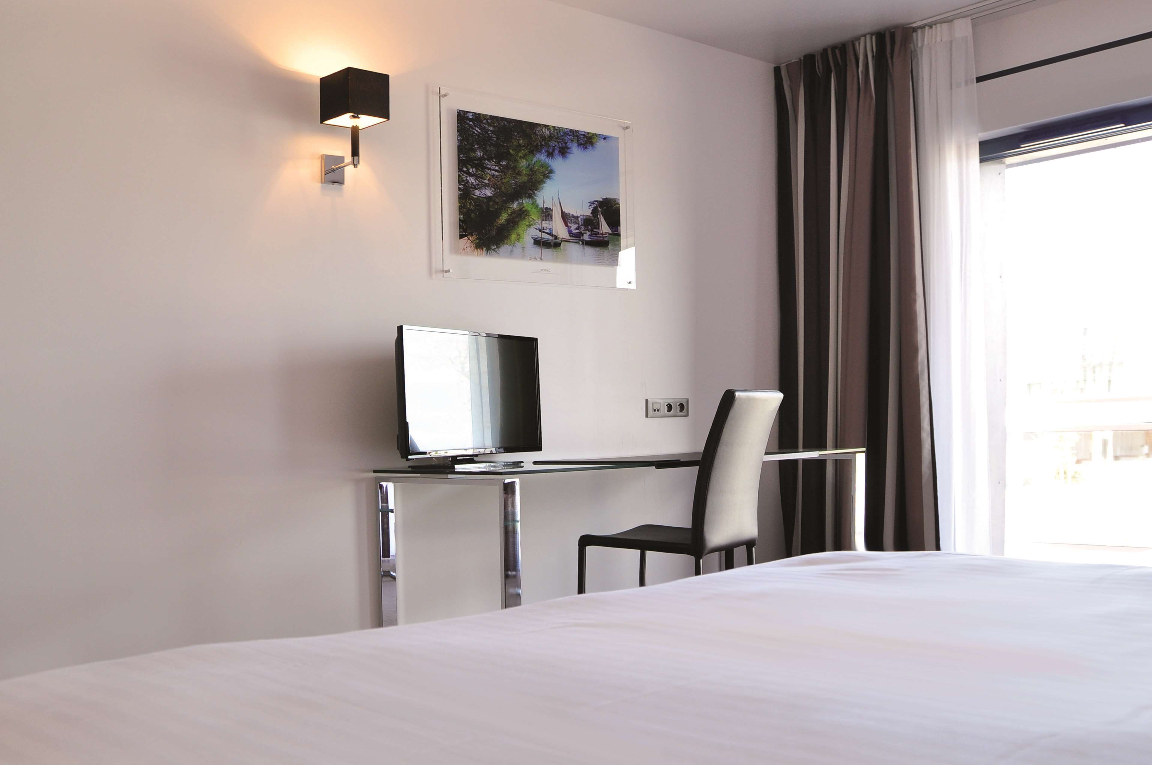 A hotel room with a bed, a desk with a computer, a chair, a wall-mounted TV, and a window with curtains.