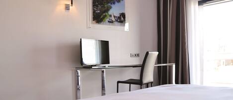A hotel room with a bed, a desk with a computer, a chair, a wall-mounted TV, and a window with curtains.