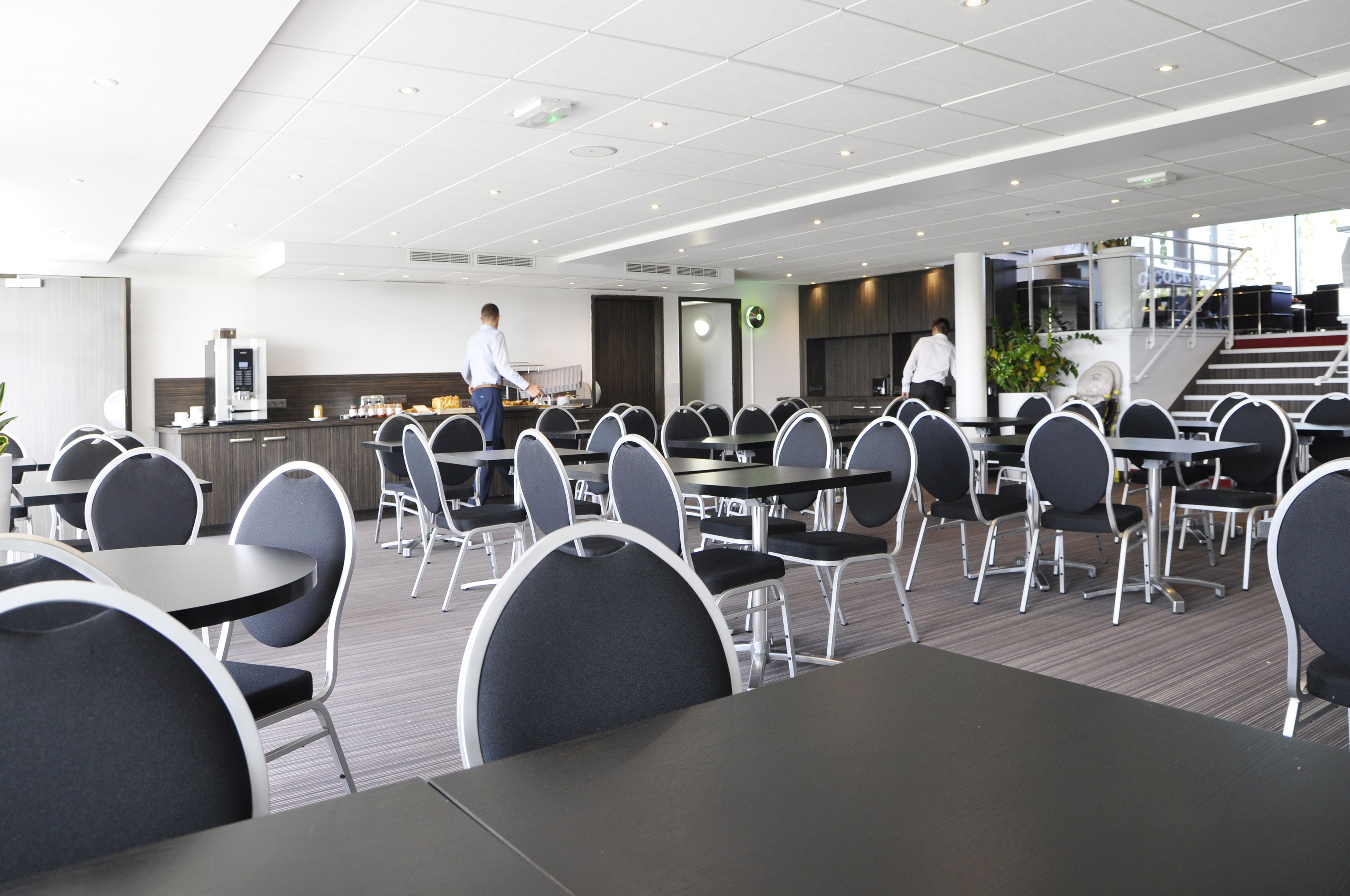 A modern dining area with round tables and black chairs, a kitchen area, and a staircase leading to an upper level.