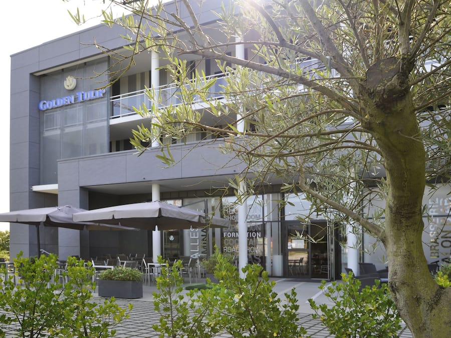 En modern byggnad med en skylt som visar texten ”Golden Tulip”. Här finns utomhusplatser med parasoller, ett träd i förgrunden och en balkong med räcken.
