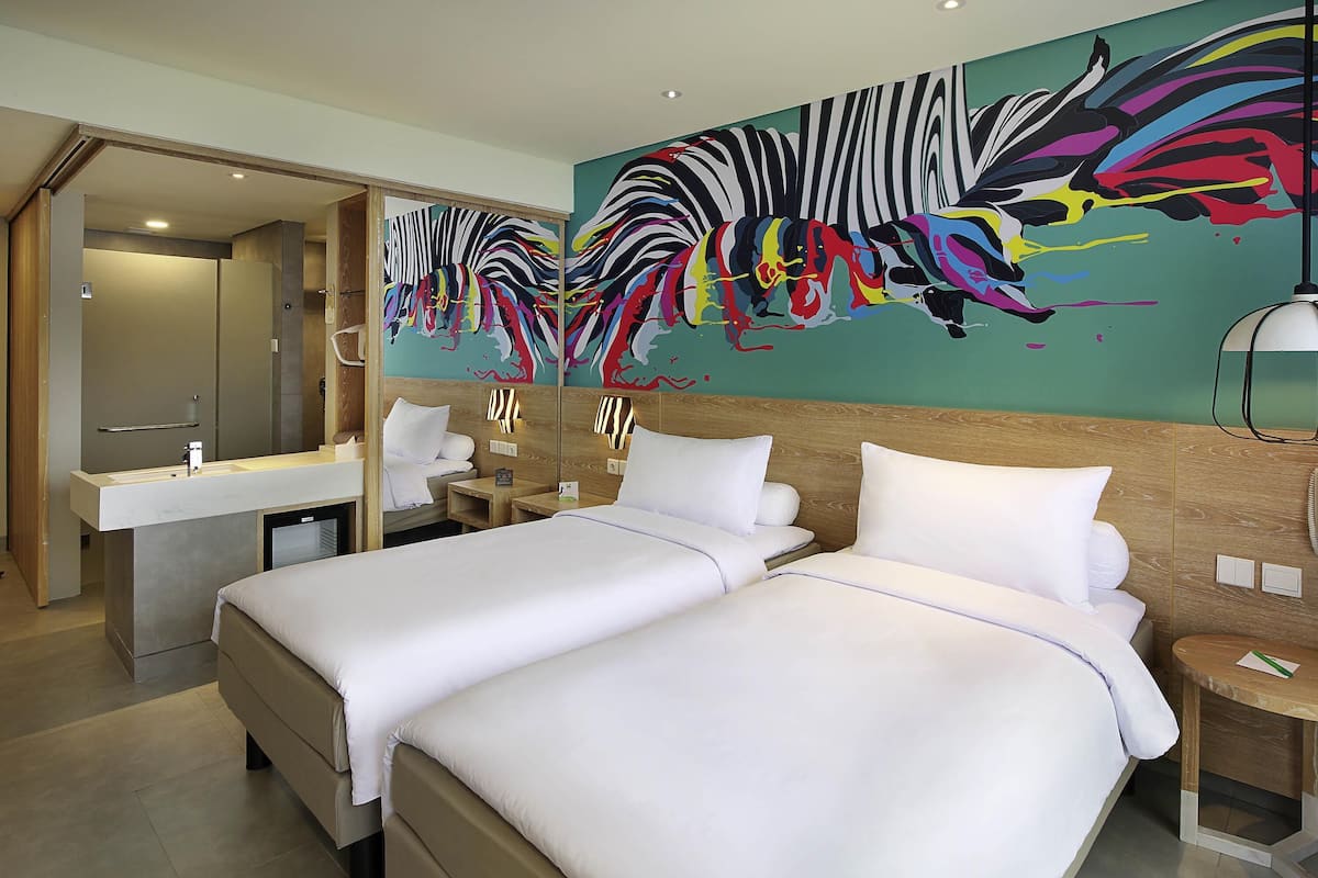 A hotel room with two beds, a bathroom with a mirror, and a colorful abstract mural on the wall.