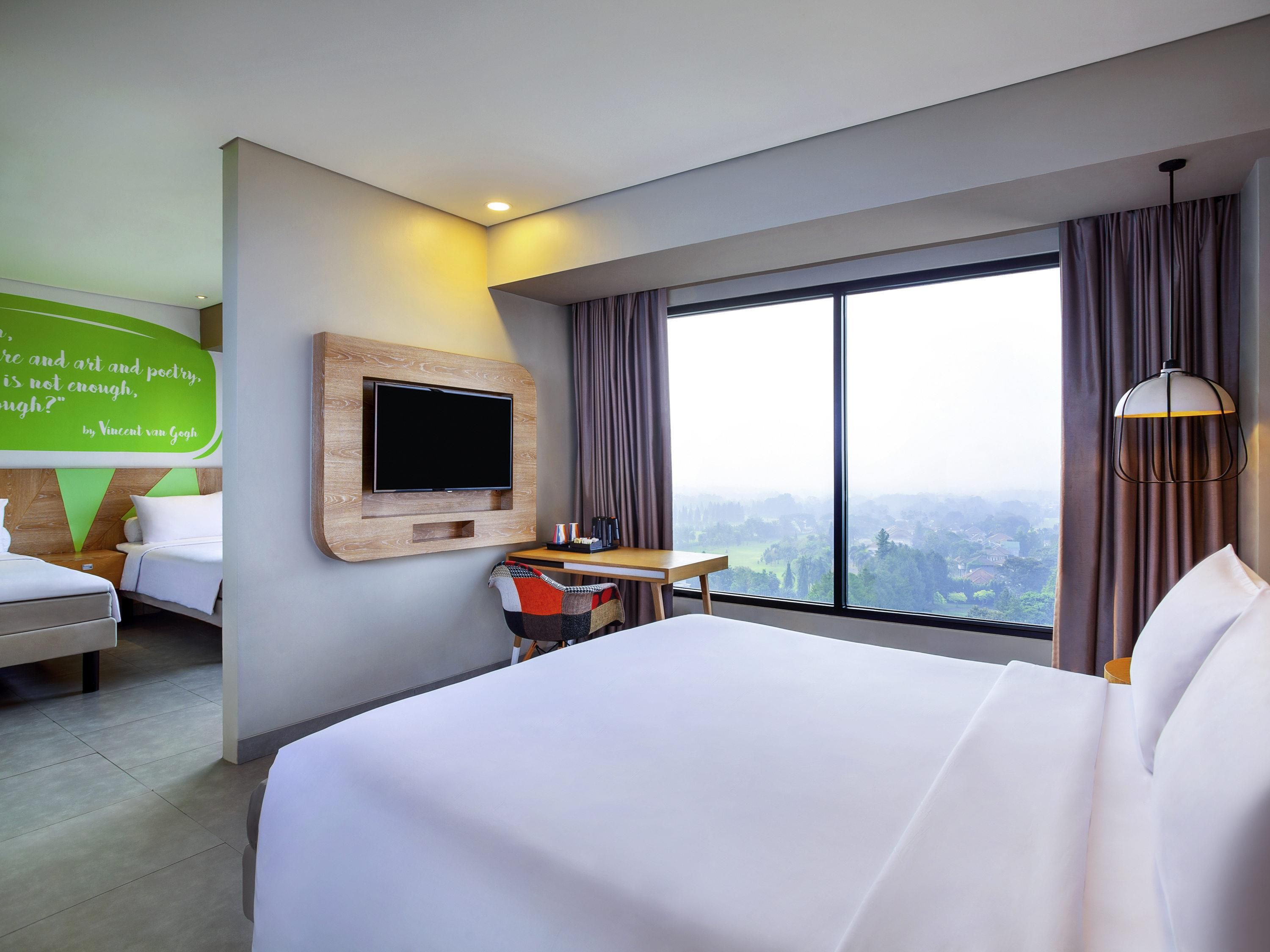 A hotel room with a large bed, a TV mounted on the wall, a desk with a chair, and a large window offering a city view.