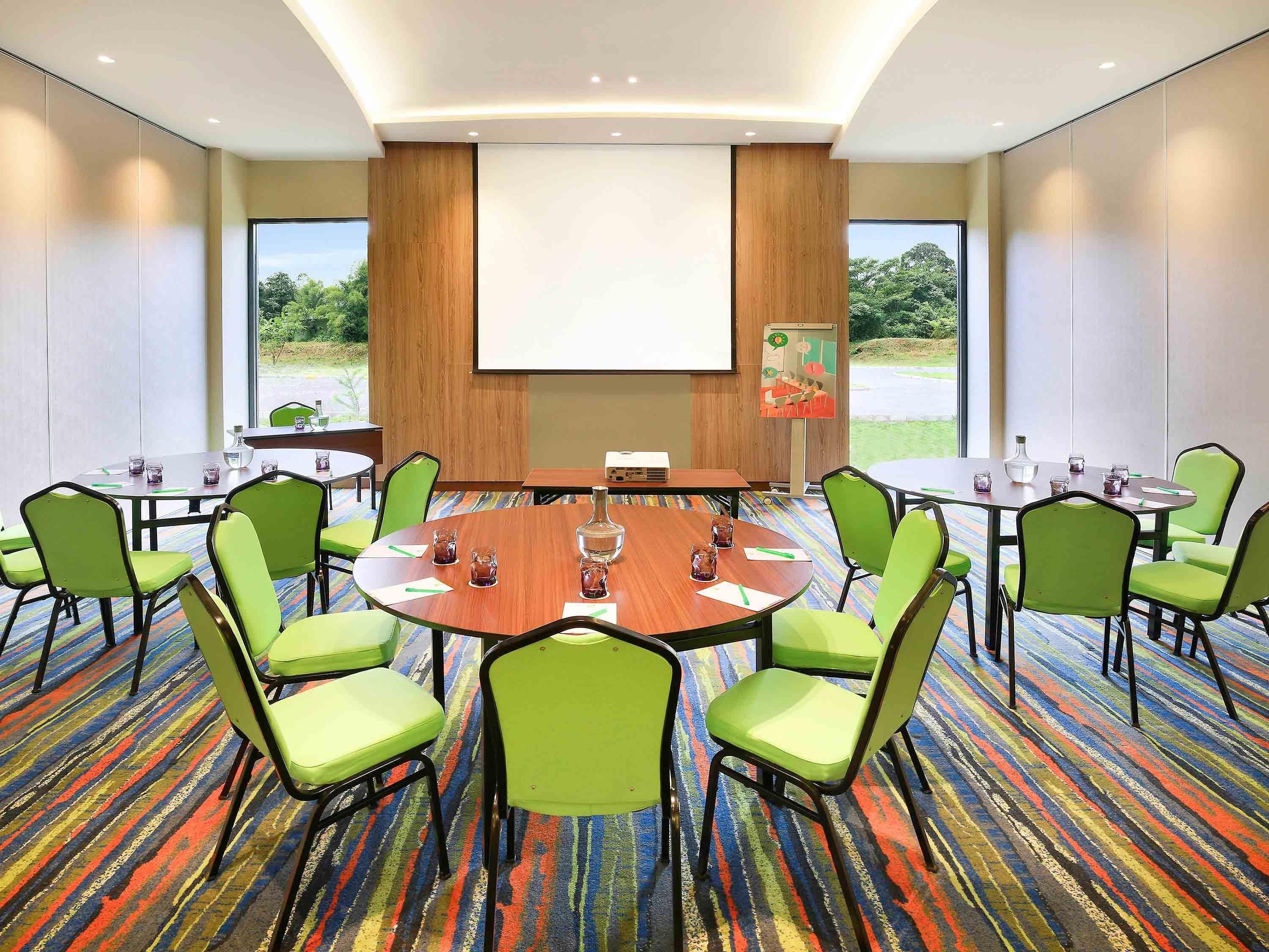 A conference room with a round table, green chairs, a projector screen, and a carpeted floor.