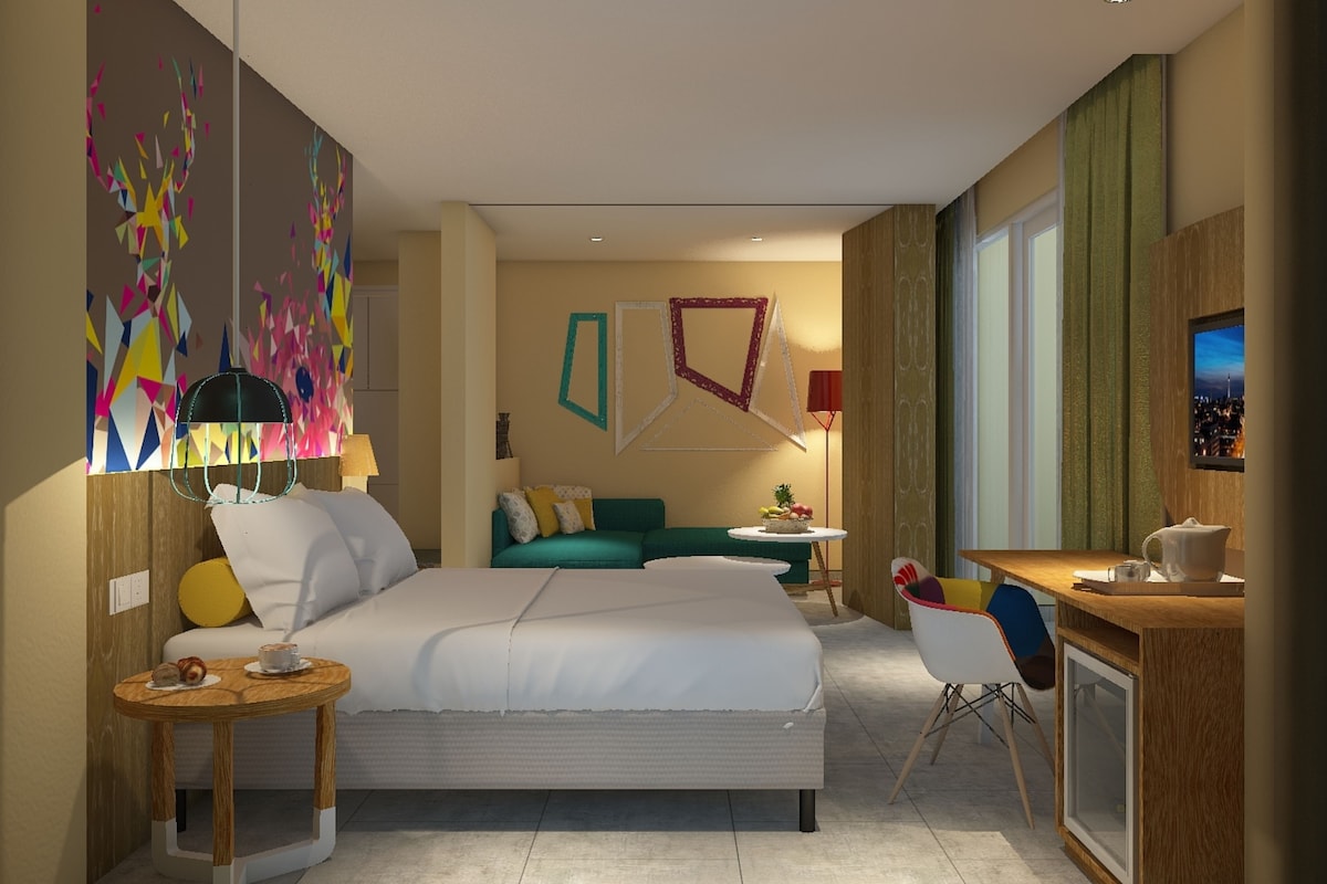 A modern hotel room with a large bed, a desk, a chair, and a small table with a lamp.