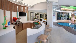 Una cocina moderna con una encimera curva, armarios de madera y un televisor integrado. Se ven taburetes coloridos y una zona de piscina al fondo.