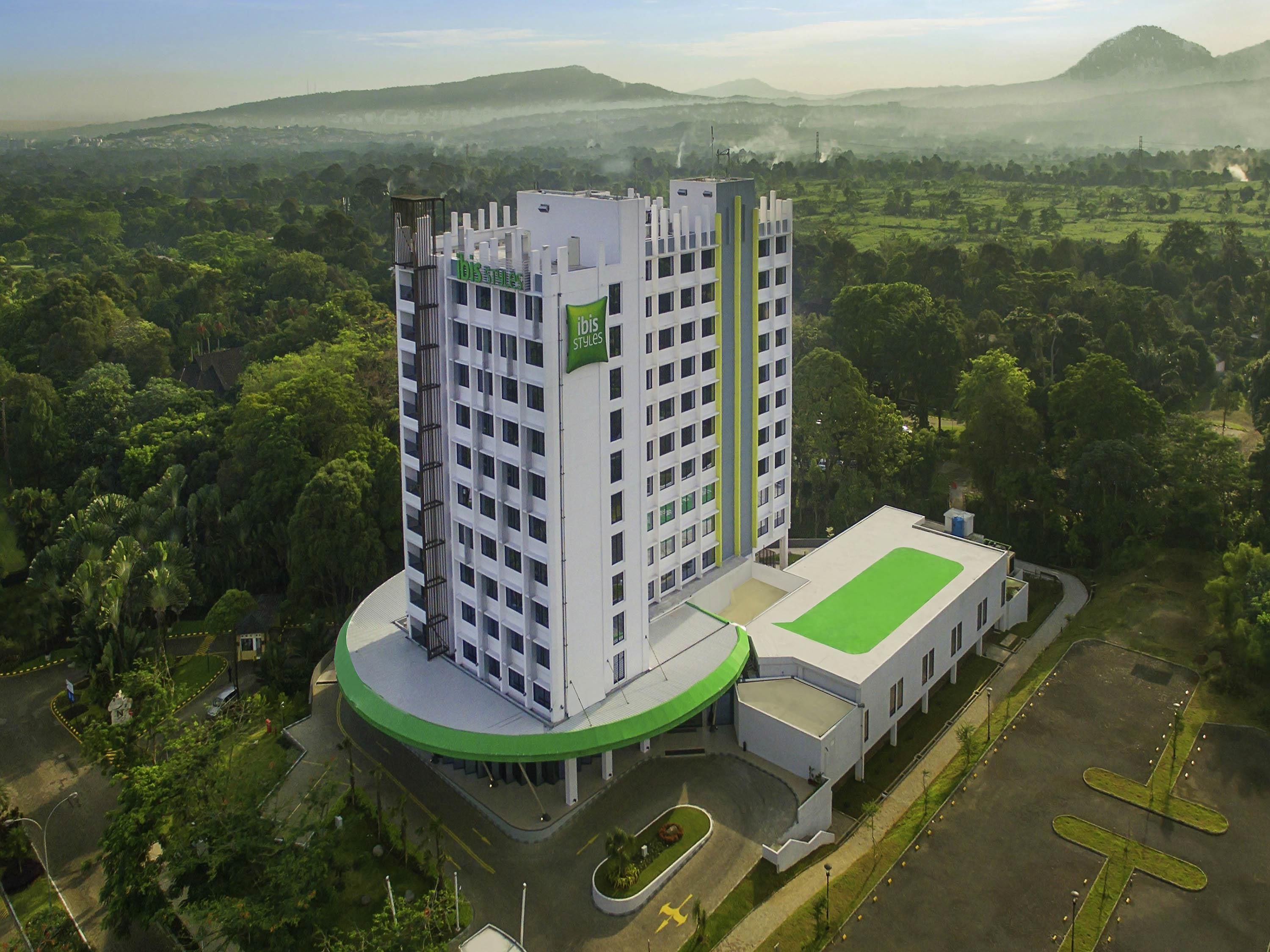 Un hotel moderno con una torre central, un cuerpo anexo de techo plano y un paisaje verde circundante.