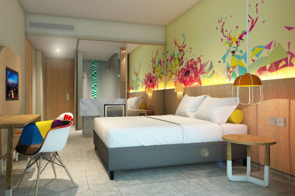 A modern hotel room with a bed, a desk, a chair, and a colorful abstract wall art.