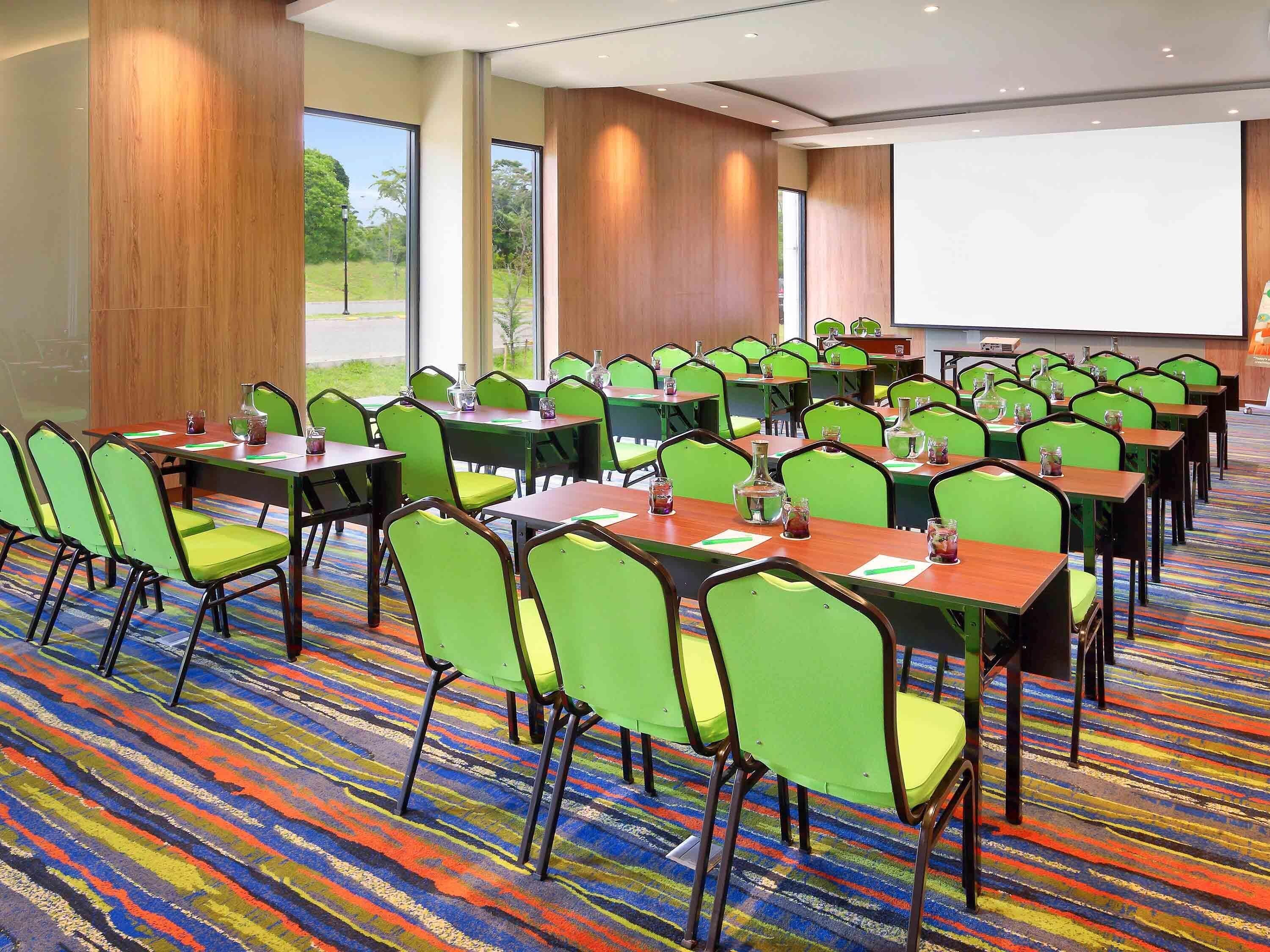 A conference room with green chairs, wooden tables, and a projector screen.