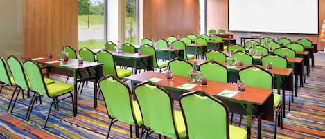 Un salón de conferencias con sillas verdes, mesas de madera y una pantalla para proyector.