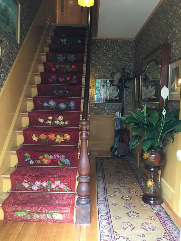A staircase with floral carpet runners, a wooden handrail, and a patterned rug leading to a room with a large mirror and a potted plant.