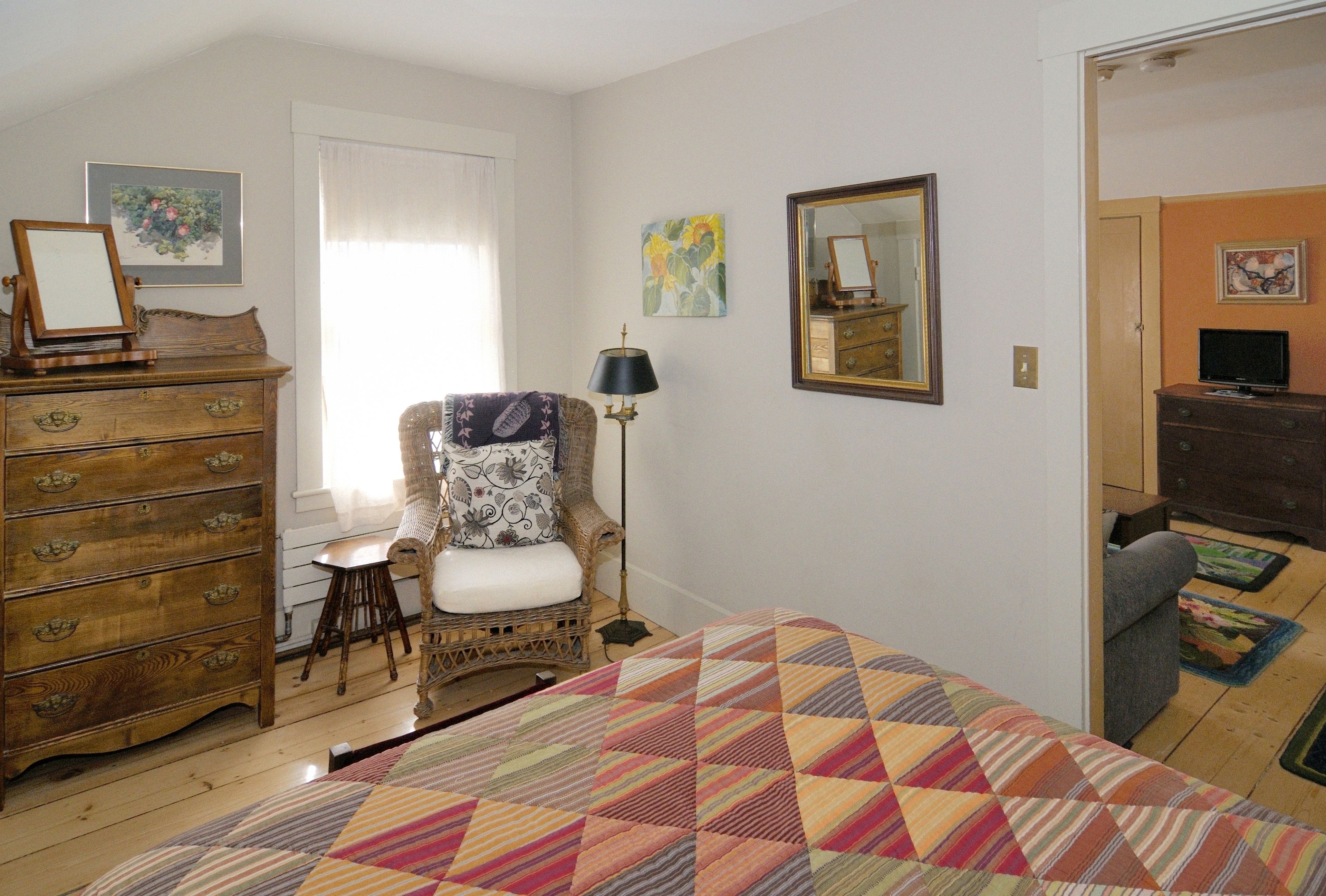 A bedroom with a wooden dresser, a chair with a cushion, a bed with a colorful quilt, a mirror, and artwork on the walls.