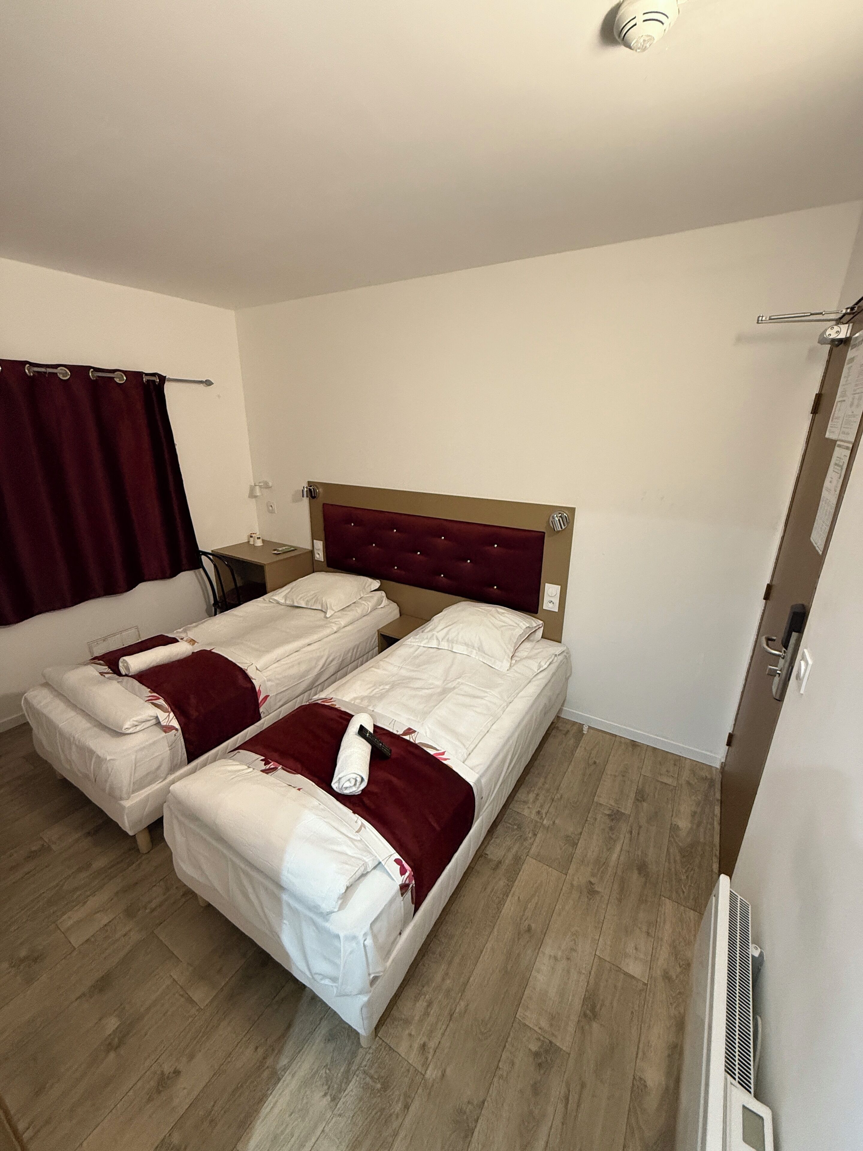 Una habitación de hotel con dos camas individuales, piso de madera, cortina roja y pared blanca.