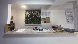 Un comptoir d'accueil avec une pancarte indiquant « RÉCEPTION, VEUILLEZ SONNER », une décoration florale fixée au mur, ainsi que divers affiches et brochures d'information.