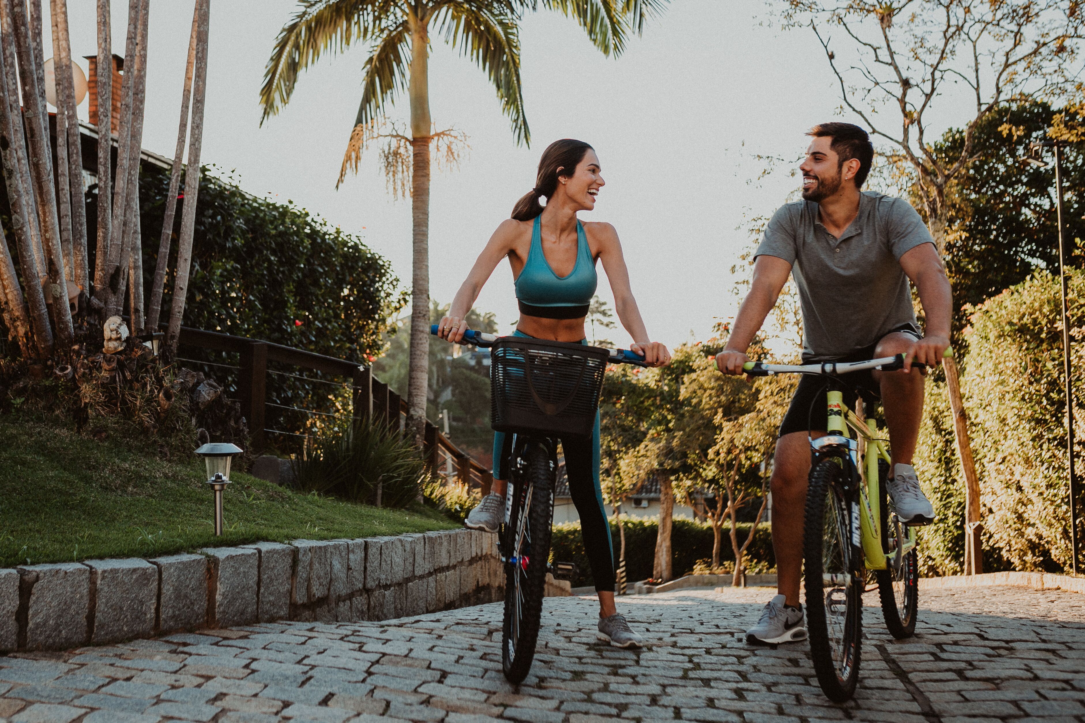 Due persone in bicicletta su un sentiero acciottolato in un parco con alberi e un lampione.