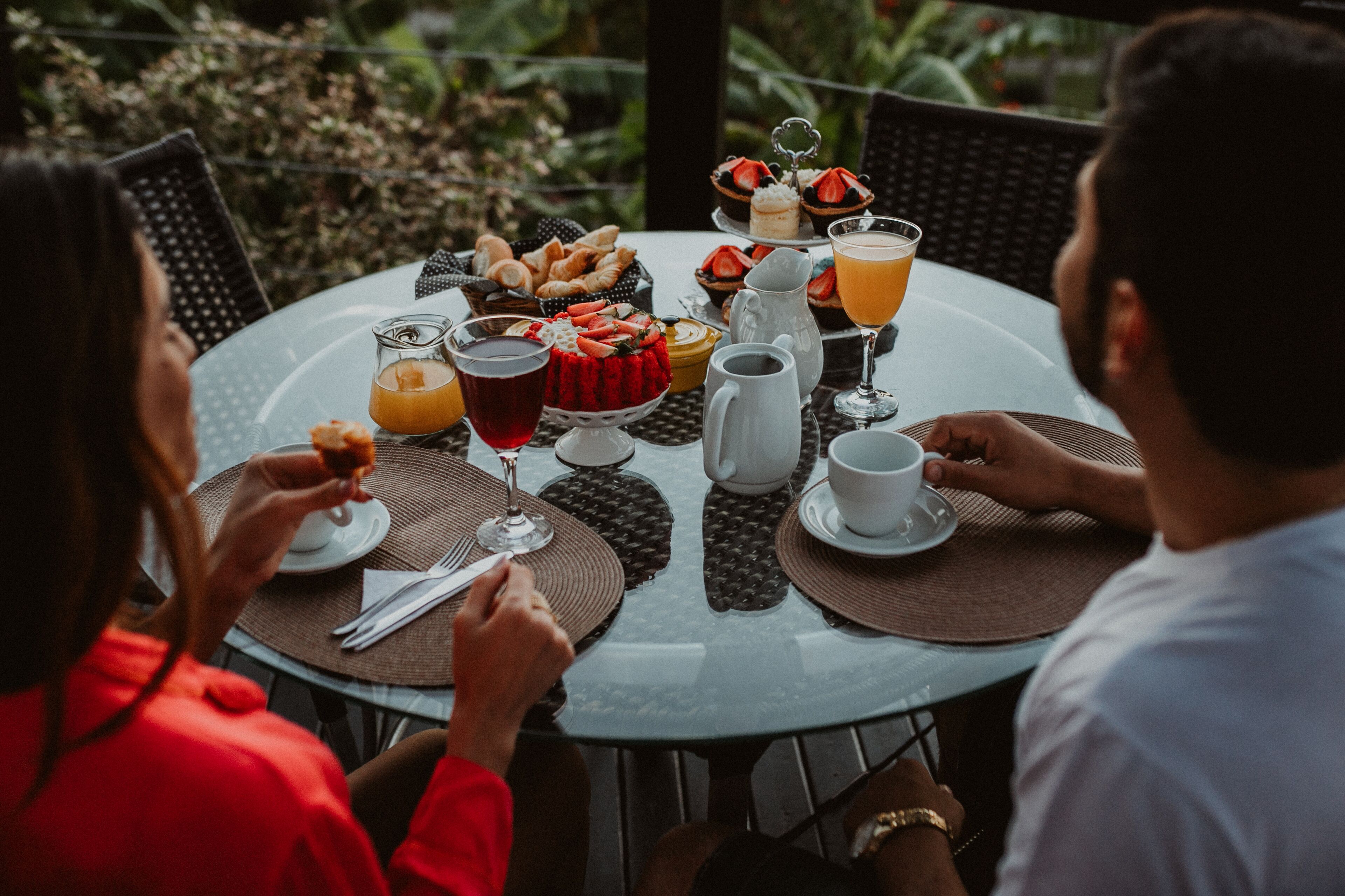 A round table with a variety of food and drinks, including pastries, fruit, and beverages, set outdoors with greenery in the background.