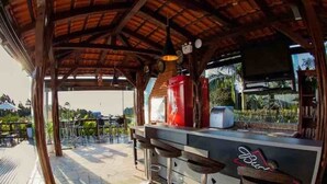 Zona de bar a l'aire lliure amb teulada de fusta, nevera de Budweiser i vista cap a la piscina.