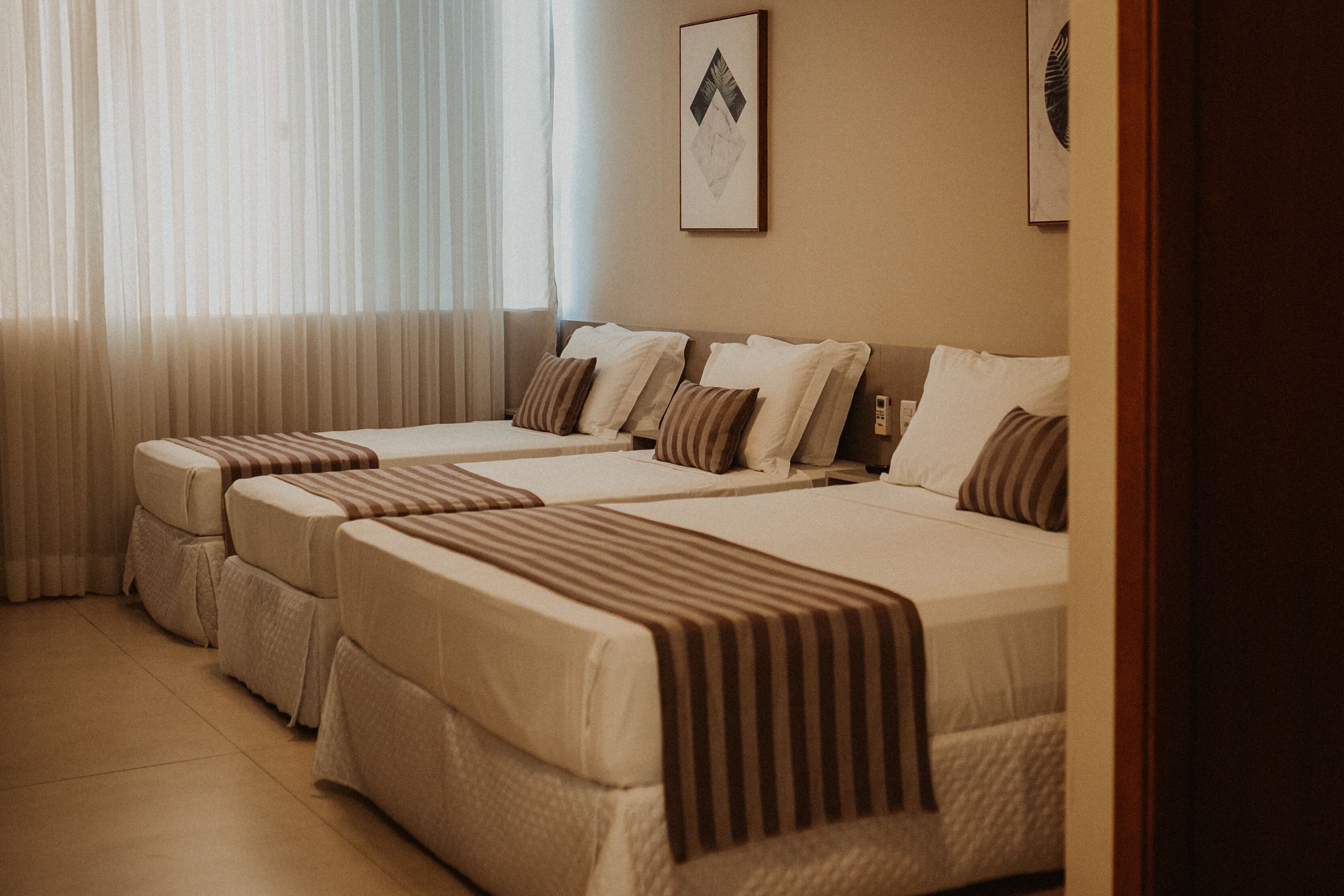 A hotel room with two beds, each with a striped blanket and pillows. There is a window with sheer curtains, and a framed picture on the wall.