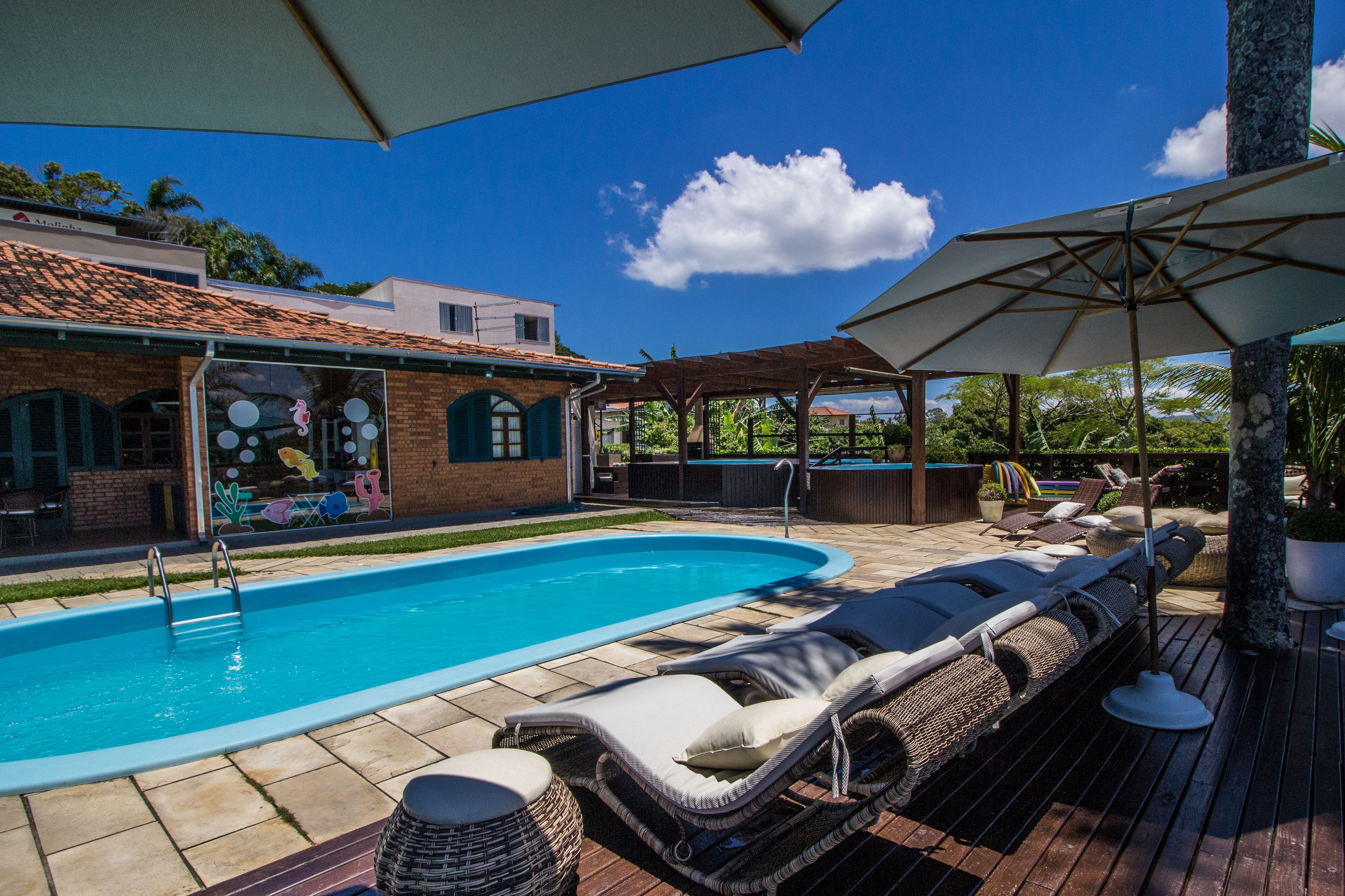 A swimming pool with lounge chairs and umbrellas, surrounded by a wooden deck and a brick building with a tiled roof.