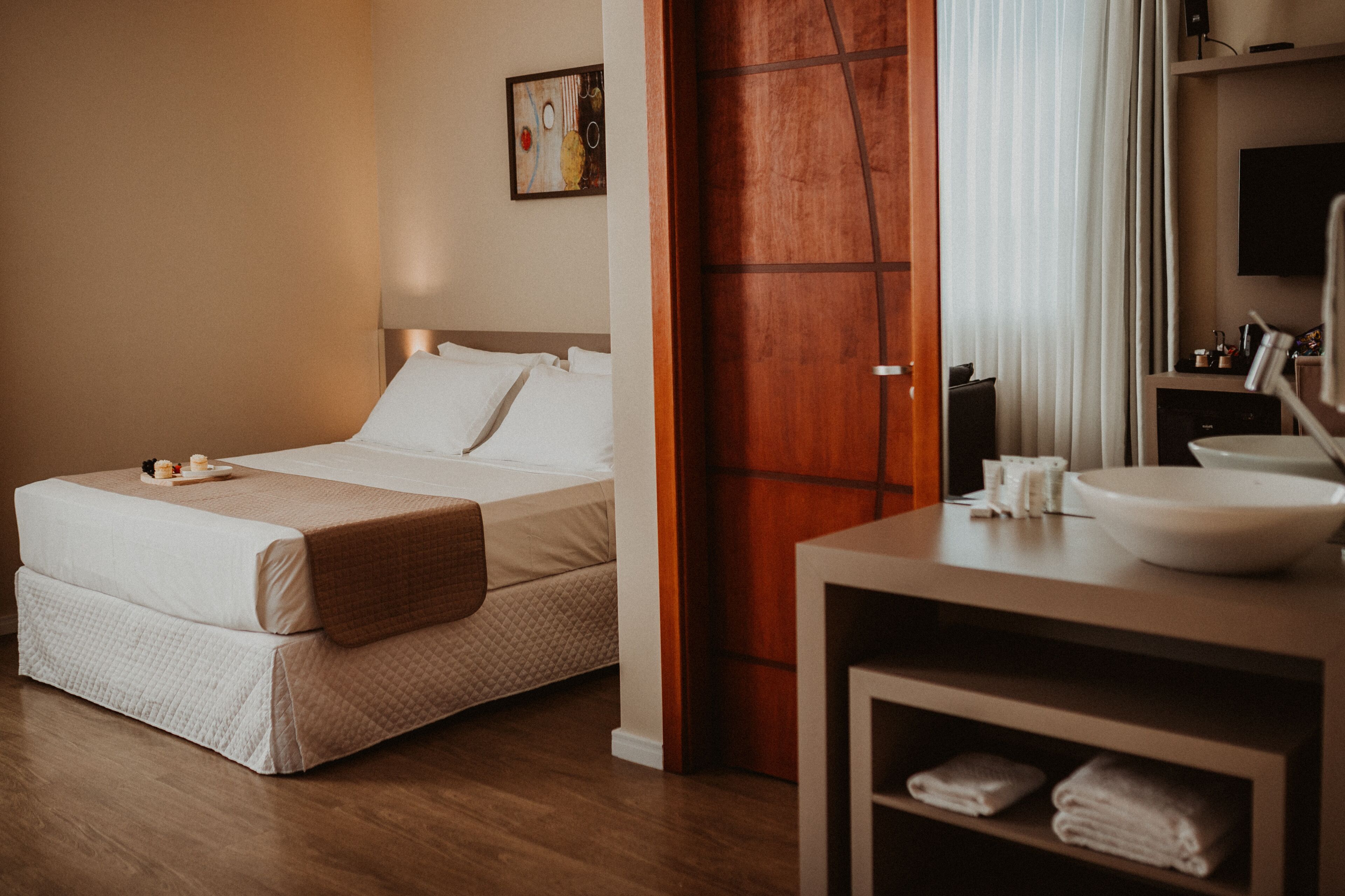 Ein Hotelzimmer mit einem Bett, einer Holztür, einem Badezimmer mit Waschbecken und Spiegel sowie einem Gemälde an der Wand.