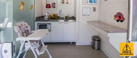 Cocina compacta con una trona, un lavamanos y una encimera con hervidor y frutero.