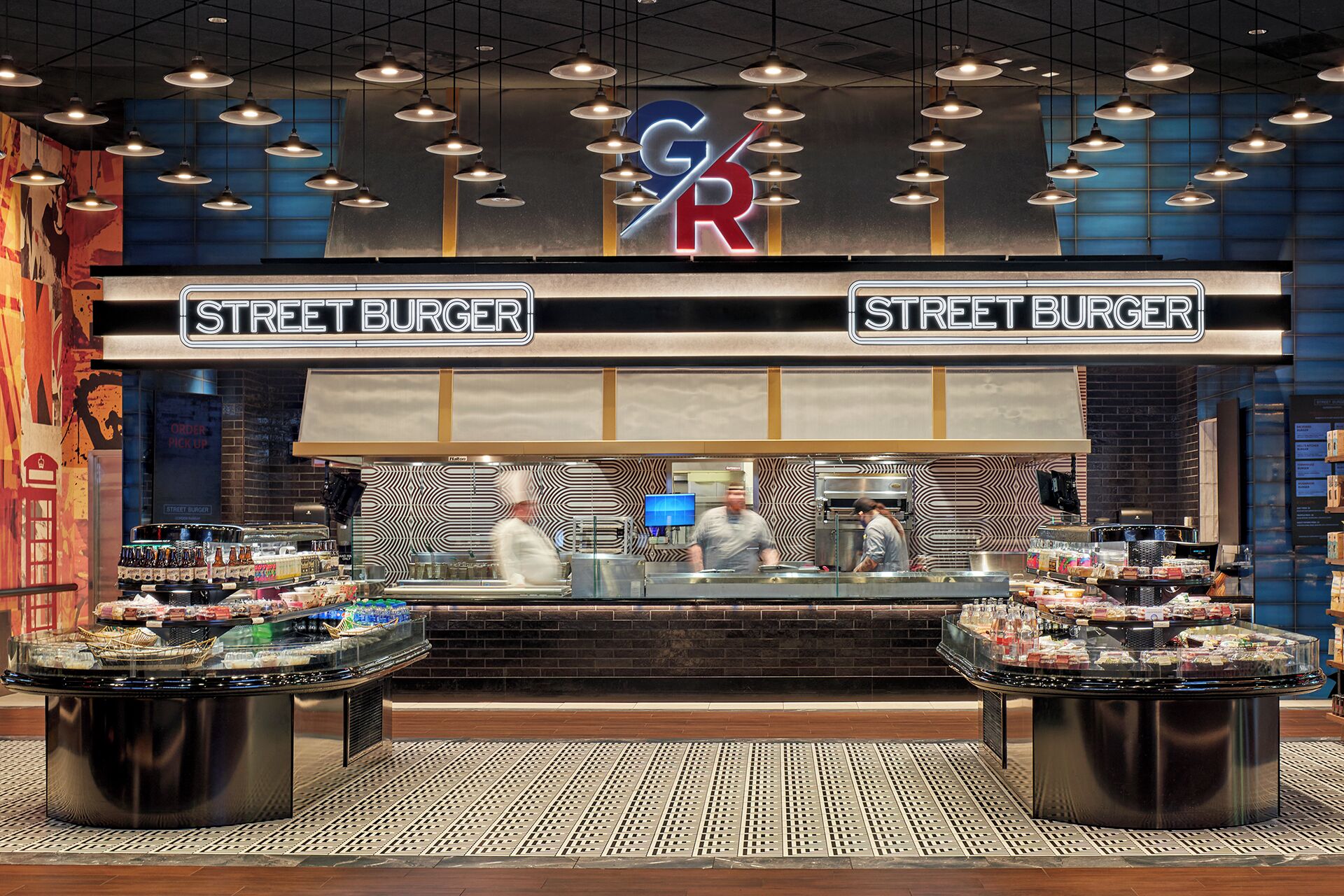 A street burger restaurant with two large display cases filled with condiments and snacks, a kitchen area with chefs, and a prominent logo above the entrance.