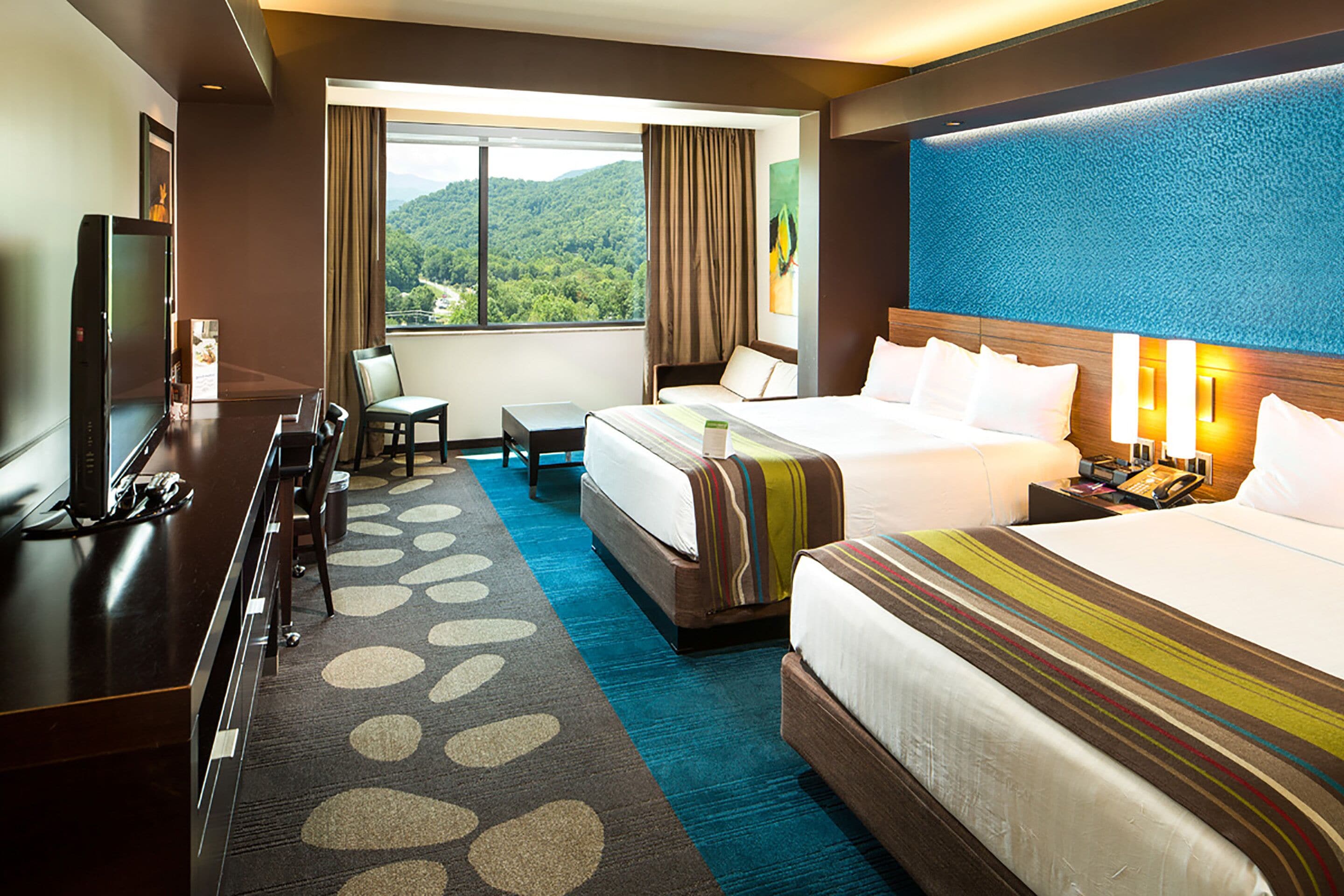 Habitación de hotel con dos camas, un escritorio, un televisor y vistas a un paisaje montañoso.