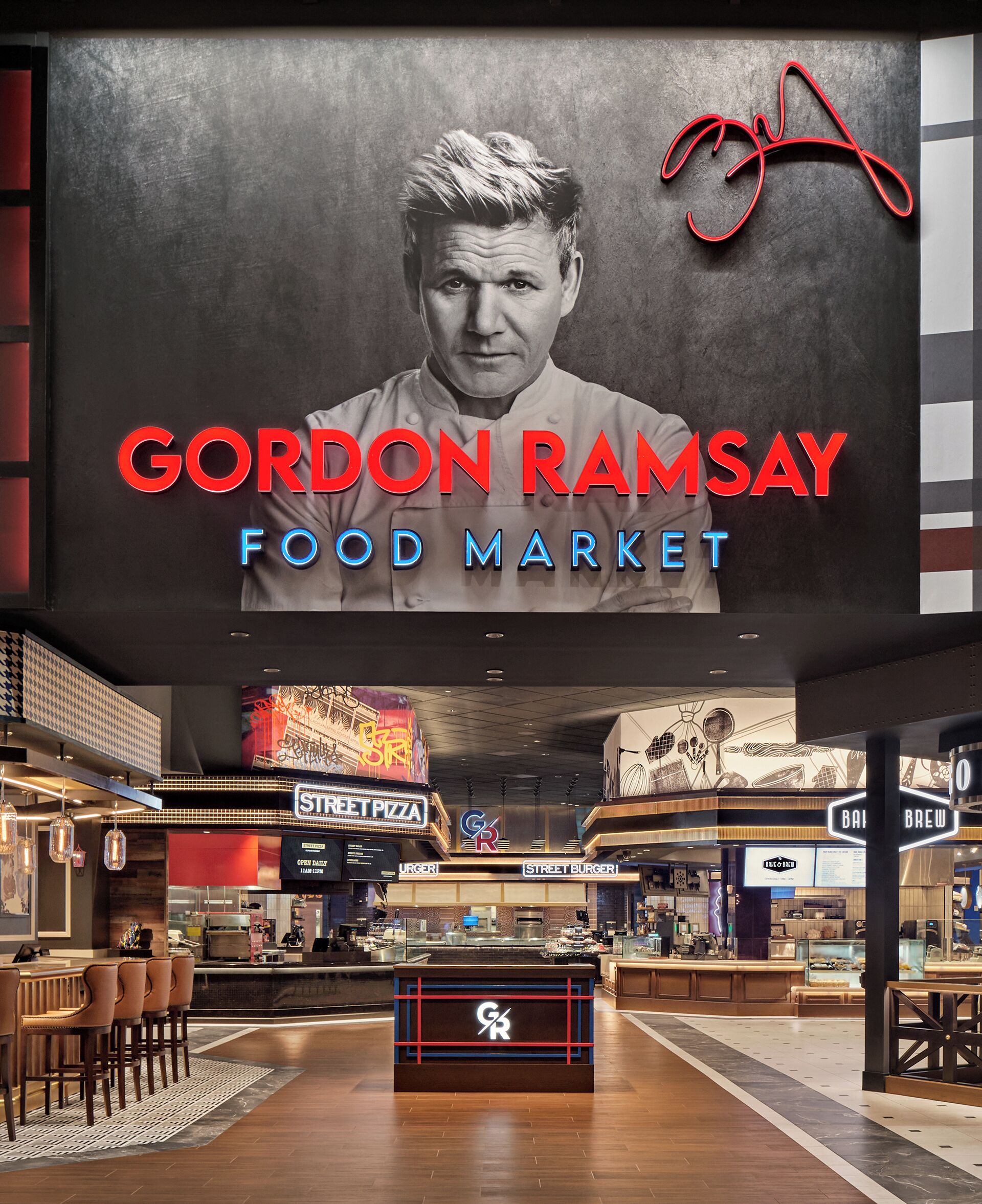 Innenansicht eines Lebensmittelmärktes mit einem auffälligen Schild für Gordon Ramsay Food Market, verschiedenen Essensständen wie Street Pizza und Street Burger, und einem Sitzbereich mit Stühlen.