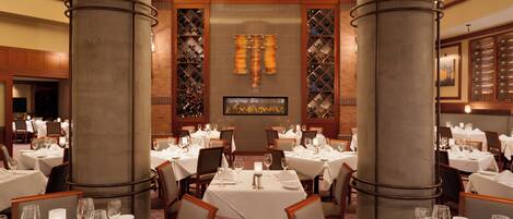 Ein Restaurant mit runden Tischen, Stühlen und einem Weinkühlschrank im Hintergrund.