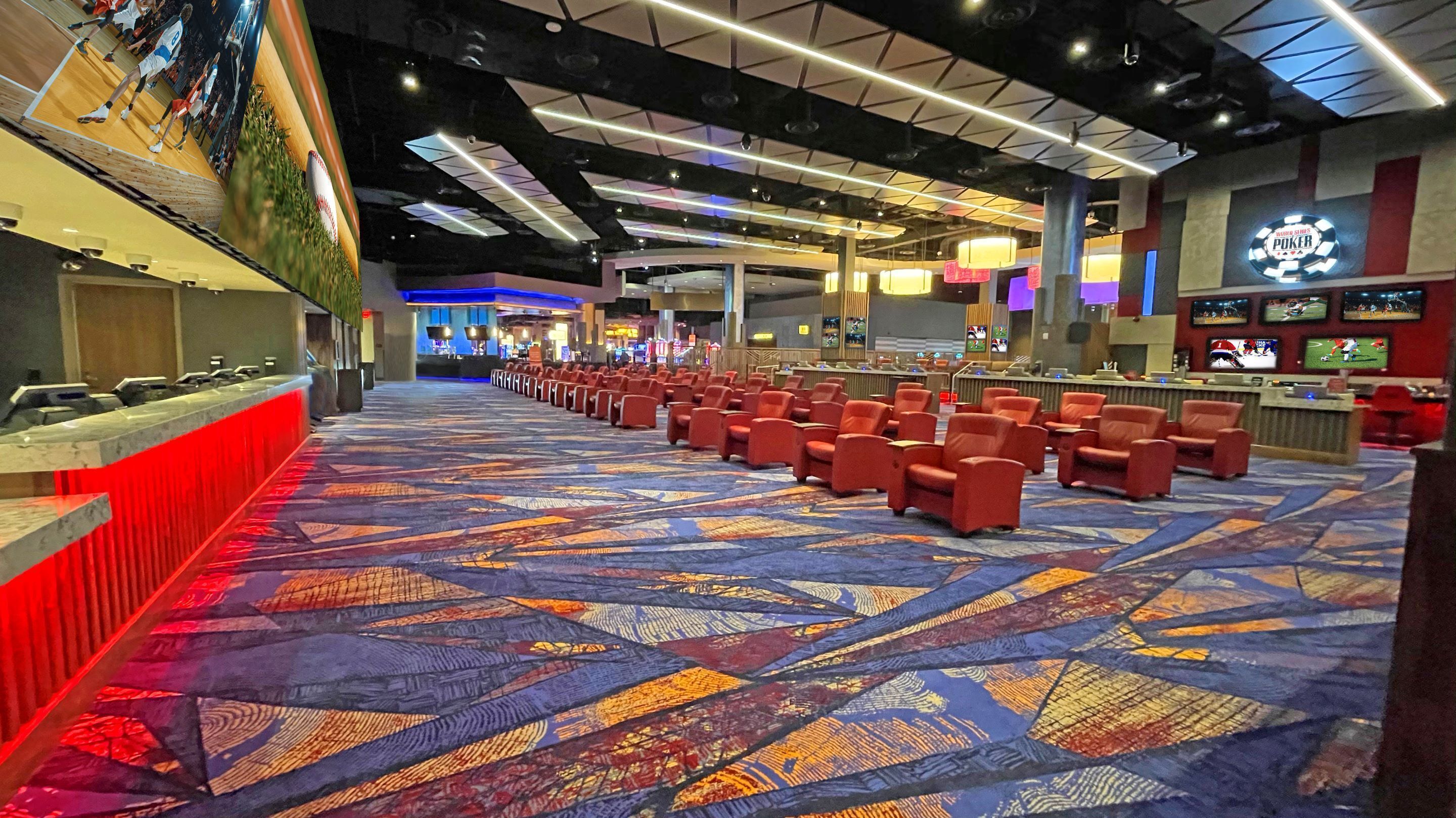 A hotel lobby with a patterned carpet, rows of red chairs, a bar area, and a large screen displaying sports.