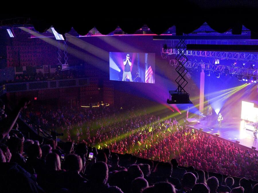 A concert with a large audience, a stage with musical instruments, and a large screen displaying the performer.