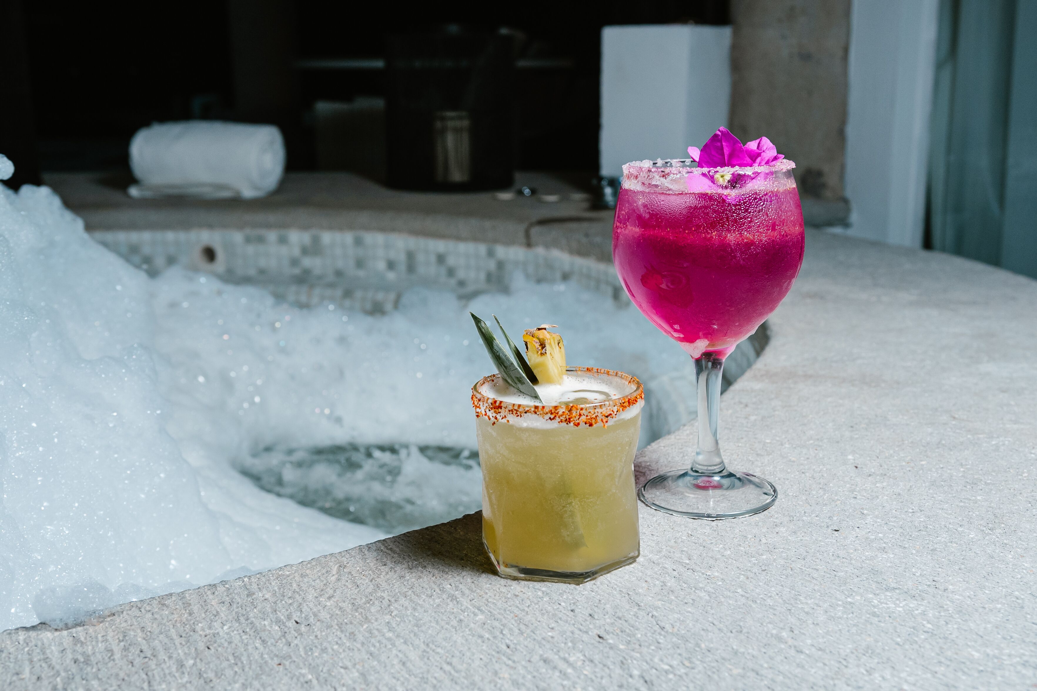 Una zona de bar con dos bebidas, una en un vaso y otra en un vaso corto, con una piscina al fondo.