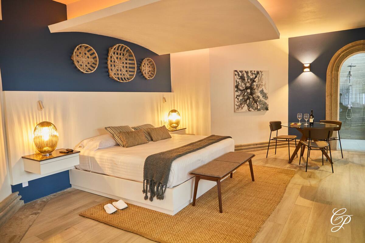 Ein Schlafzimmer mit Bett, Nachttischen, einer Bank, einem Essbereich mit Stühlen und Blick ins Freie.