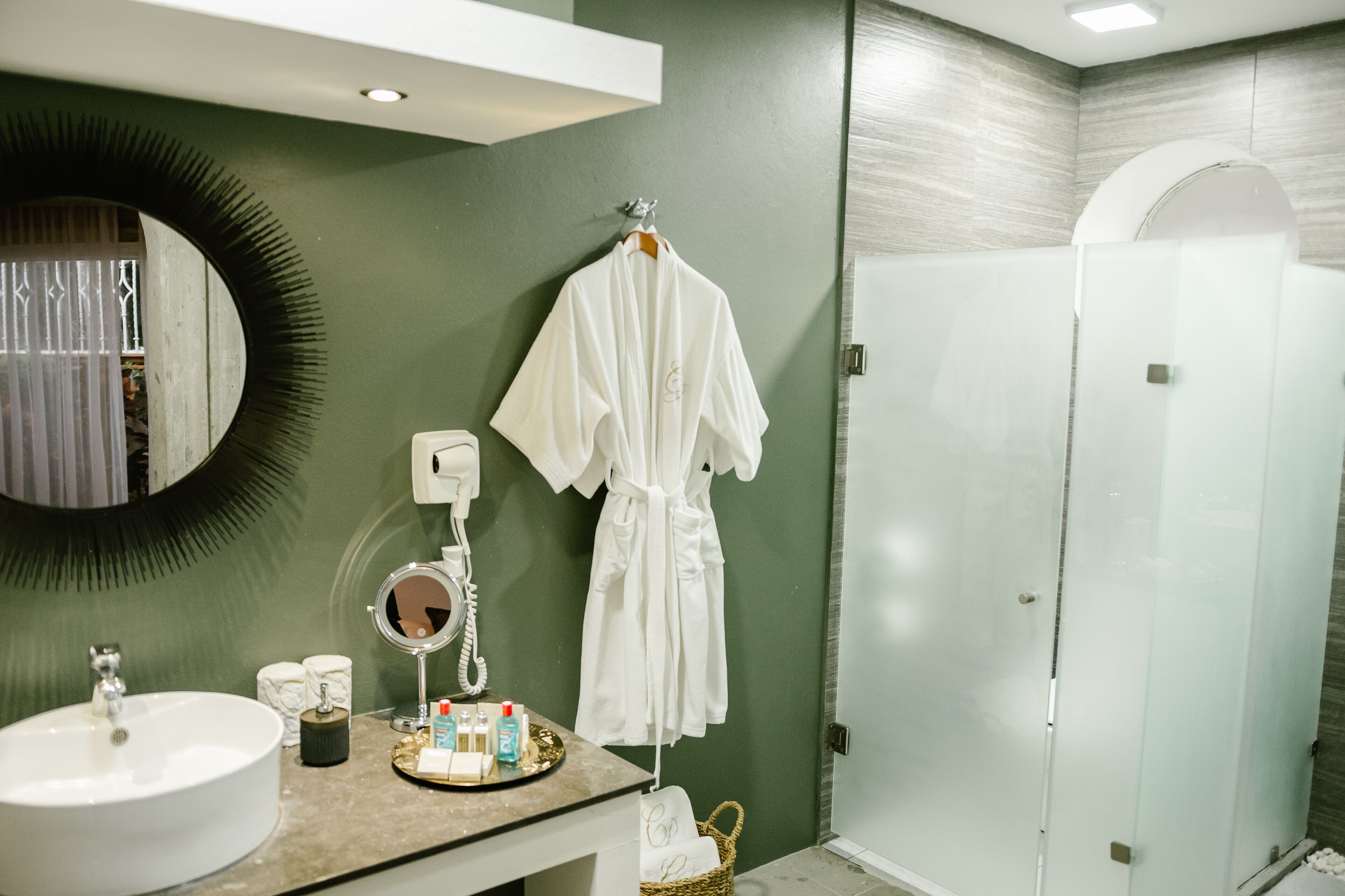 Une salle de bain avec un miroir rond, un lavabo, des articles de toilette, une robe suspendue et une douche séparée par une paroi en verre.