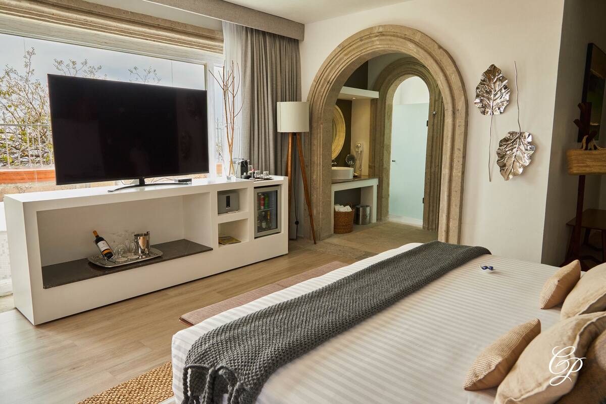 Ein modernes Hotelzimmer mit einem großen Flachbildfernseher, einem Konsolentisch mit einer Weinflasche und Gläsern, einem Bett mit grauer Decke und einem Spiegel mit dekorativen Blättern.
