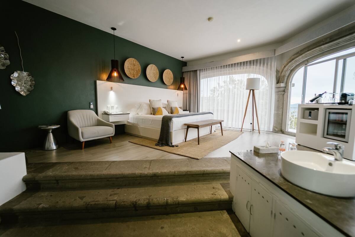Ein modernes Schlafzimmer mit Bett, Nachttisch, Lampe und Blick ins Freie durch große Fenster.