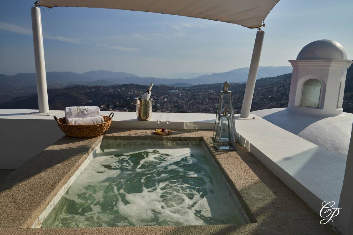 Eine Dachterrasse mit Whirlpool, einem Korbwicker-Korb, einem Glas Wein und Blick auf Stadt und Berge.