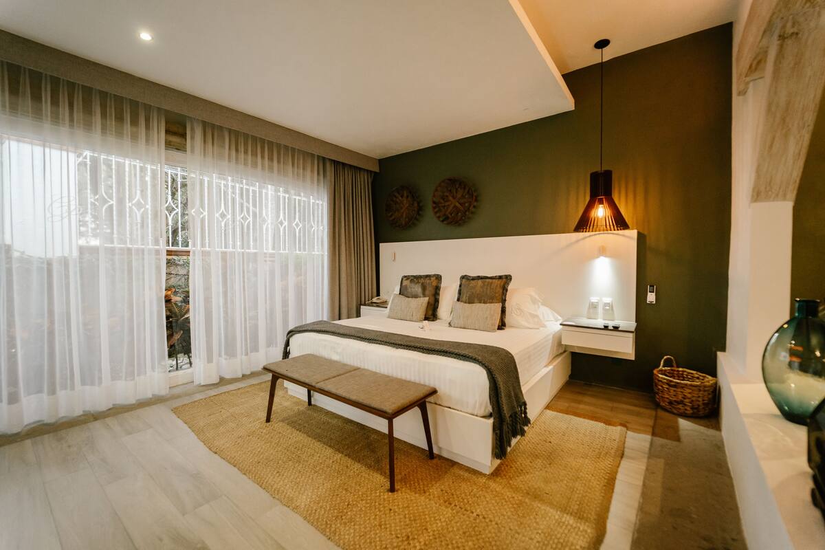 Ein modernes Schlafzimmer mit einem großen Bett, Nachttischen, einer Bank und Blick durch transparente Vorhänge.