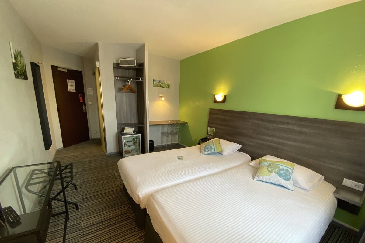 Quarto de hotel com duas camas, uma mesa de escritório e uma cadeira. Há um cabideiro e uma pequena mesa com abajur.