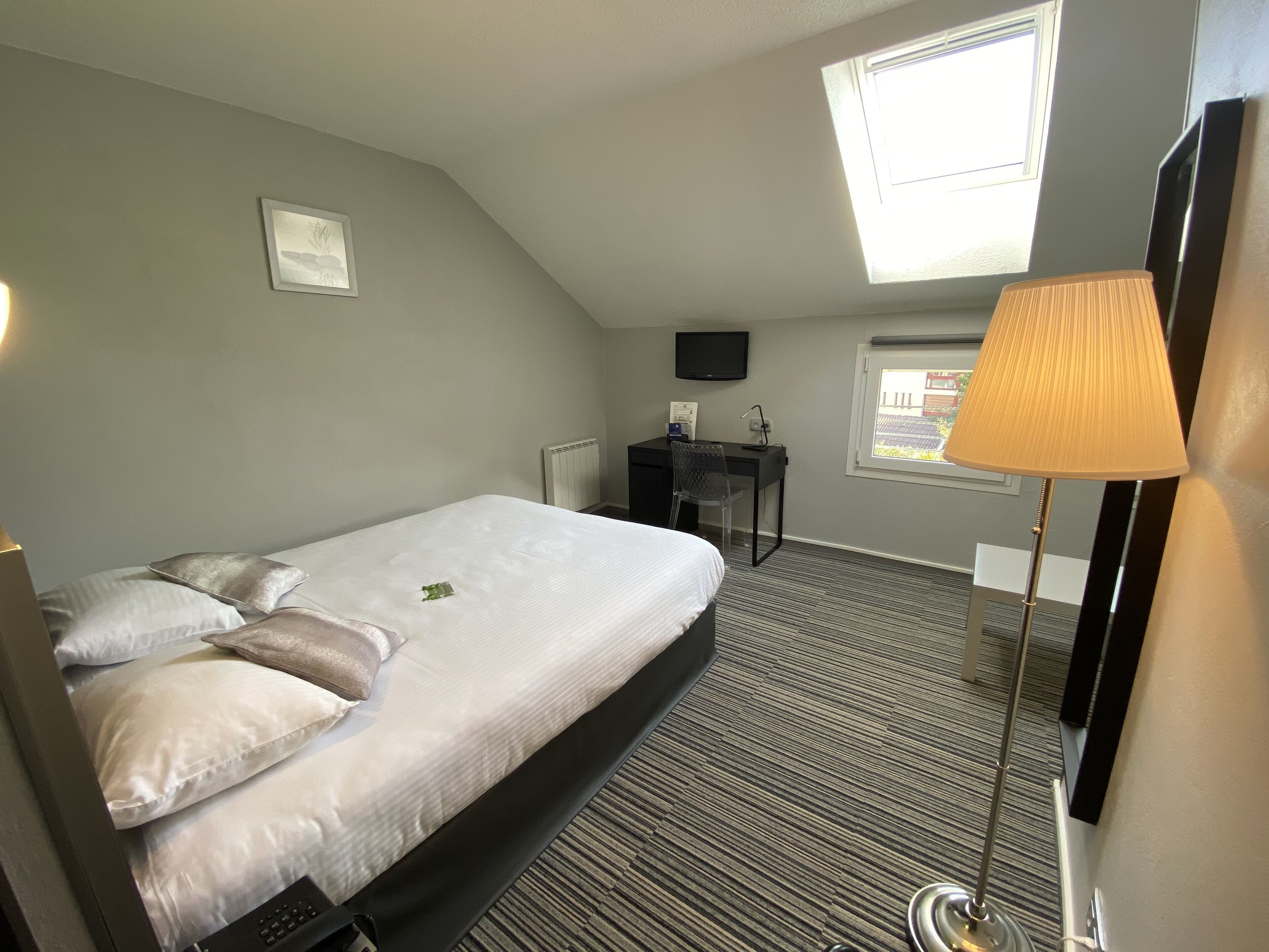 A hotel room with a bed, a desk, a chair, a TV, a window, and a lamp.